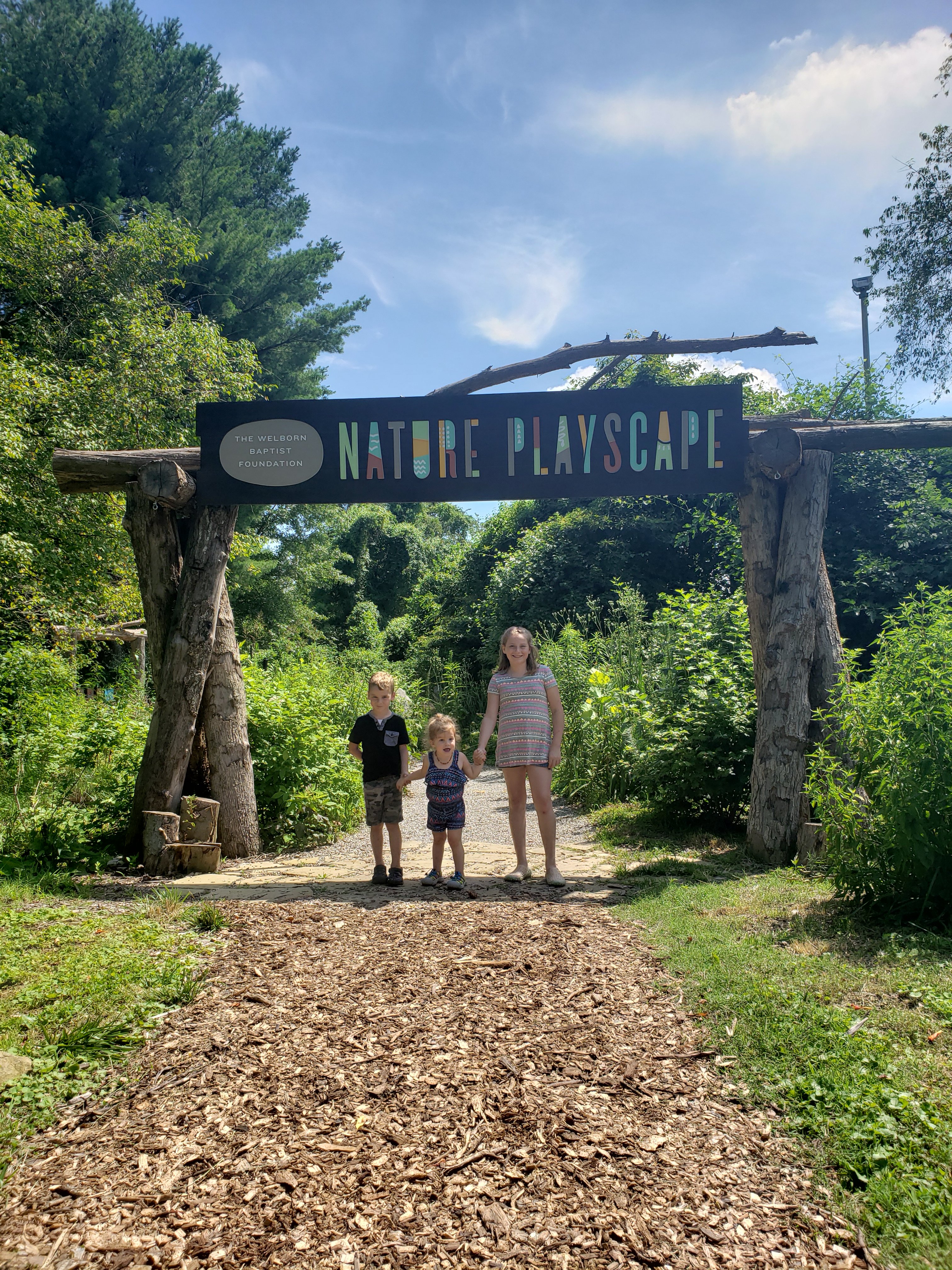 Photo of three kids holding hands under a sign that reads "Nature Playscape" at Wesselman Woods in Evansville, IN  Things to do with kids in Evansville Indiana. Kids activities in Evansville 