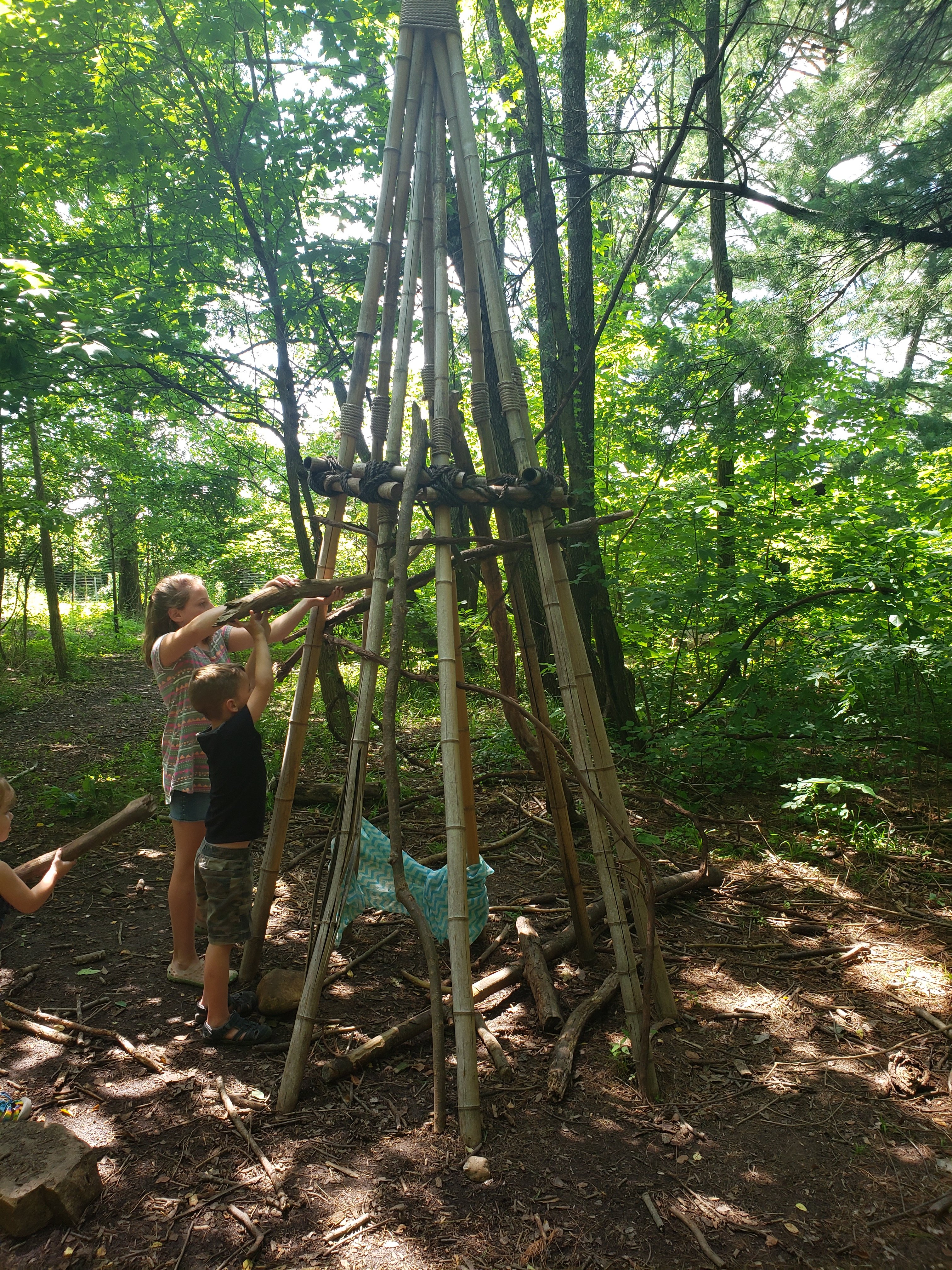 Two kids, a girl and a boy, building a teepee using natural materials, pictured here large sticks, in Wesselman Woods Nature Playscape in Evansville, Indiana  Things to do with kids in Evansville Indiana. Kids activities in Evansville 