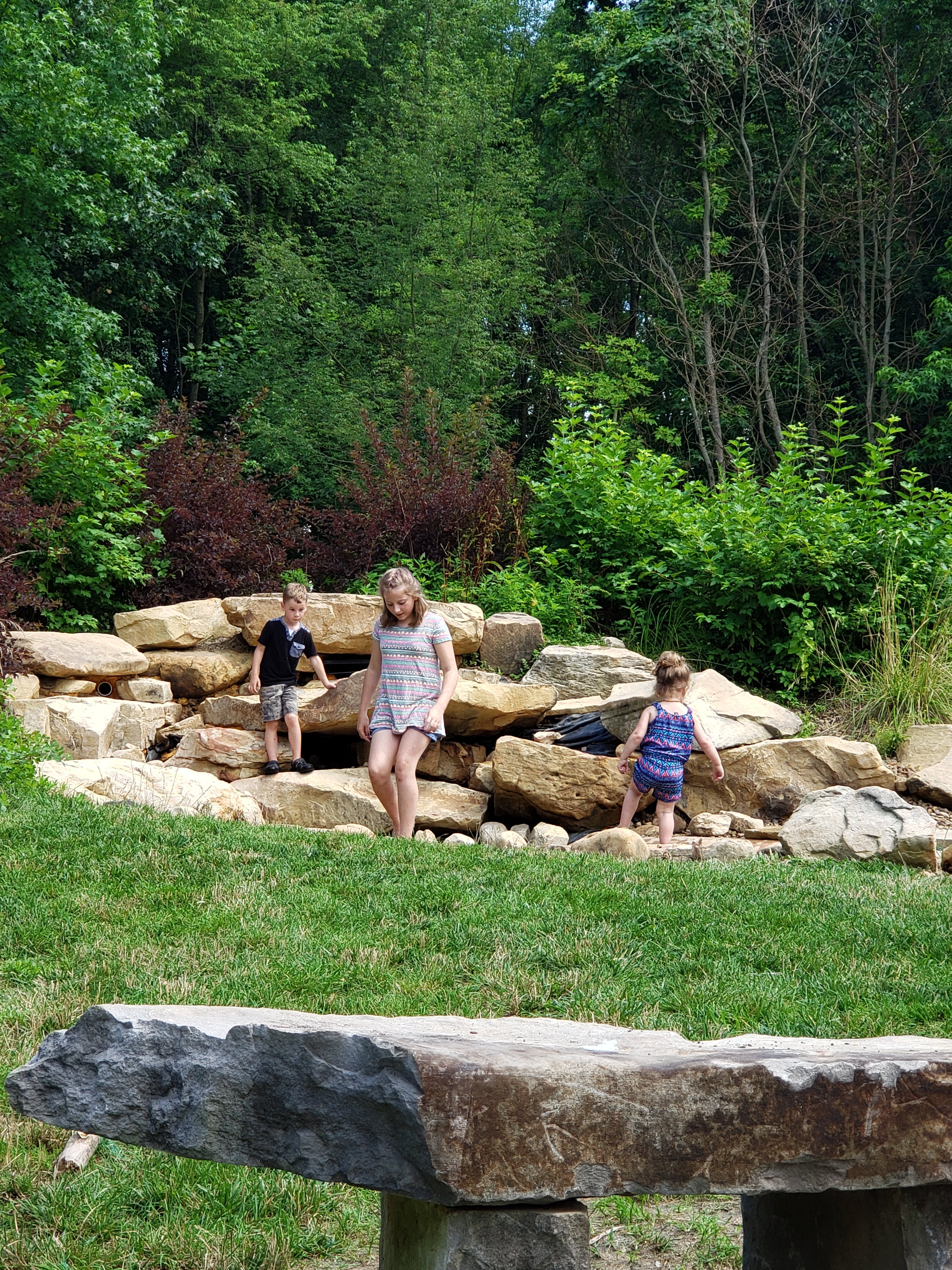 Three kids playing on large rocks at Wesselman Woods nature playscape for kids in Evansville, Indiana  Things to do with kids in Evansville Indiana. Kids activities in Evansville 