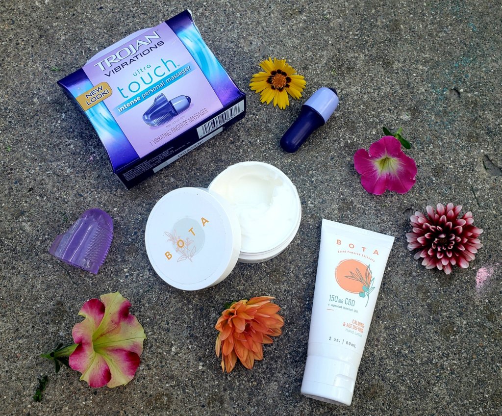 flowers, intense personal massager, BOTA muscle cream, and hand lotion laid out on cement surface