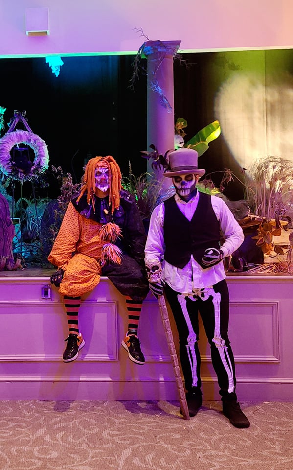 two people dressed up, one as a fancy skeleton and one a orange clown in front of a stage with more Halloween themed decor