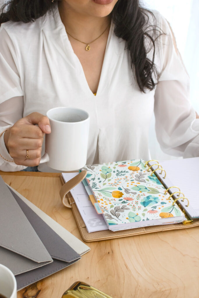 Brunette woman in a white shirt holding a coffee mug at a table with a planner 