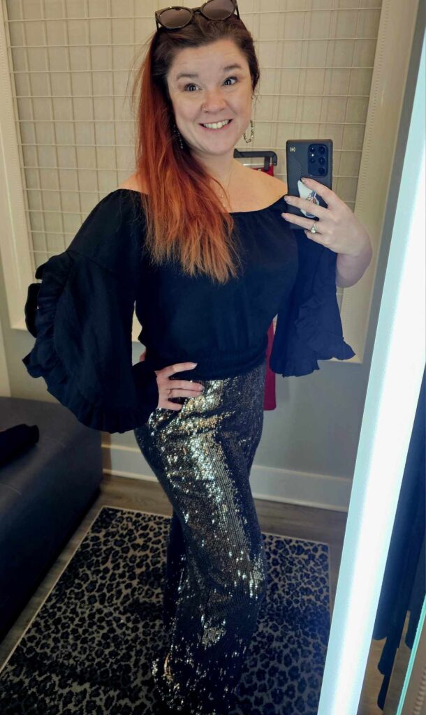 Woman with bright red hair taking a selfie of herself in the mirror wearing a flowy black top and sequin pants