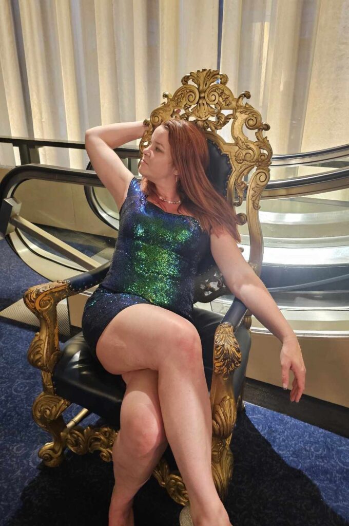 Red headed woman in a emerald green sequin dress looking to the side while sitting in a throne like gold chair with her legs crossed embrace your true self and embrace being too much