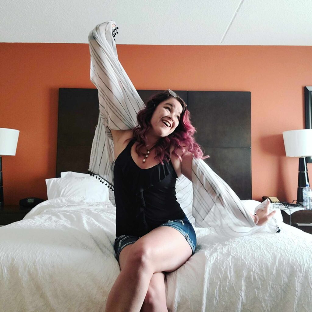 Woman with magenta hair looking happy with life as she gazes towards a window, wearing shorts and a flowy cover up with a black shirt embrace your true self and embrace being too much