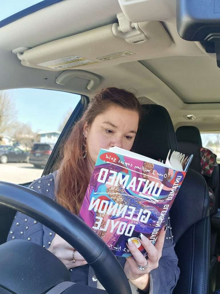 Woman sitting in a car reading Untamed by Glennon Doyle as an empowerment book for working moms