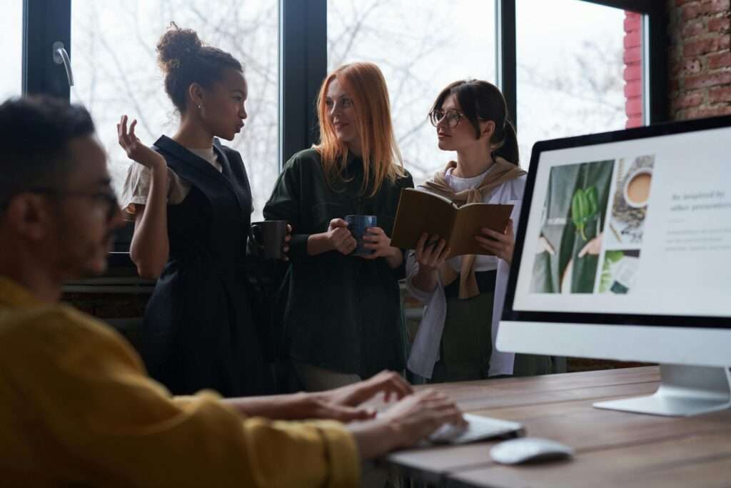 Three women chatting in an office setting in front of a window with a man working on a computer sitting in the foreground