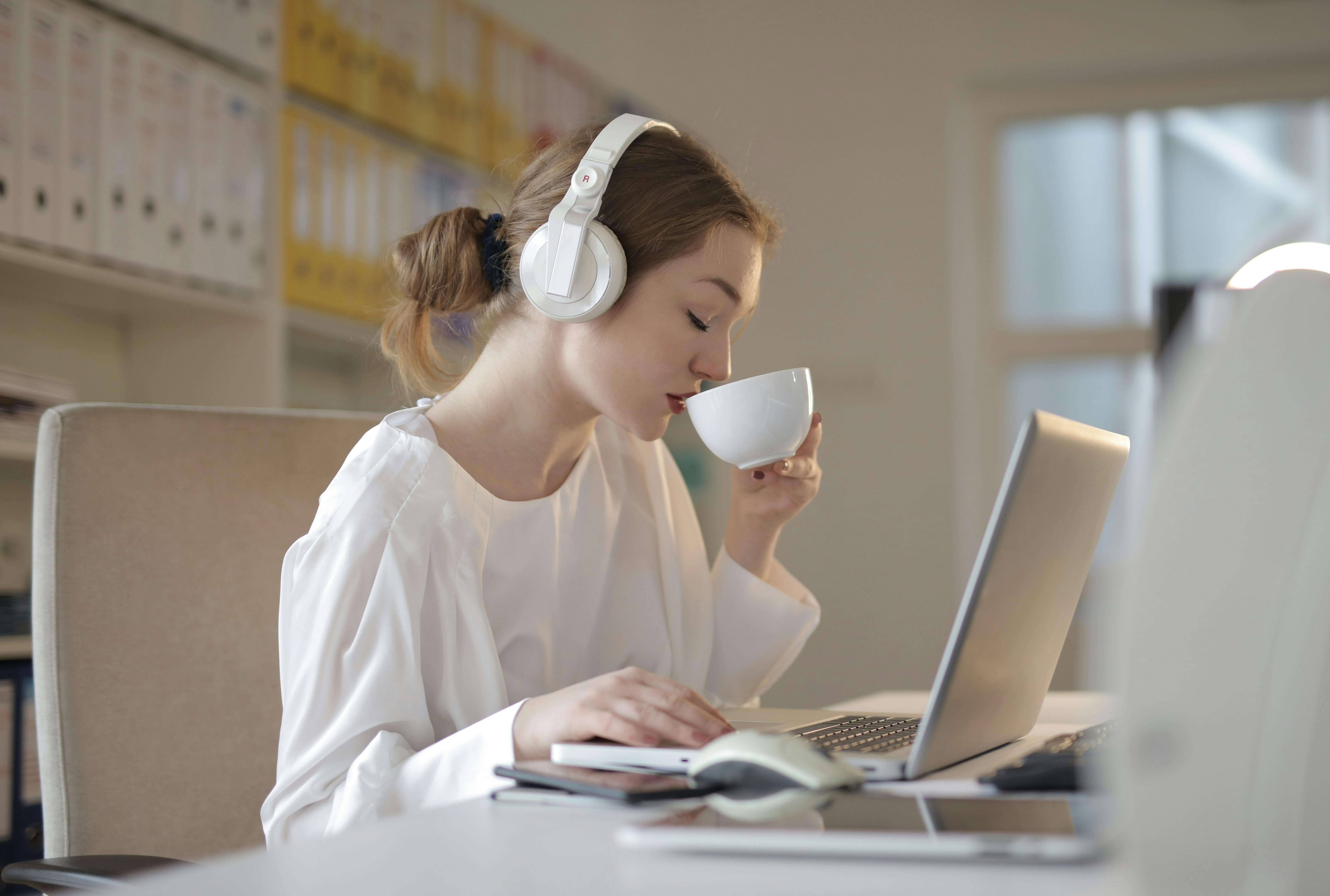 Woman with her hair in a bun sipping from a coffee mug with her eyes closed and headphones on in front of a computer practicing the power of positive influence 